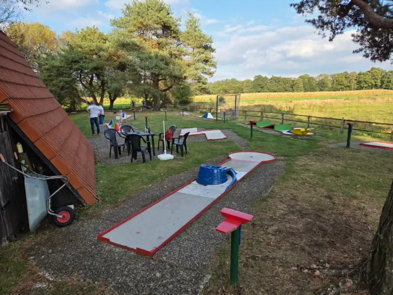 Blick über die Minigolfanlage mit der Holzhütte, Sitzgruppe und Bahnen unter hohen Kiefern