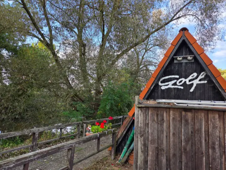 Die Golf-Hütte mit rotem Spitzdach und Schriftzug „Golf“, daneben der Weg am Wasser mit Geranien
