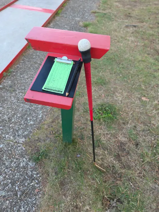 Papier-Scorekarte mit Bleistift auf einem Bahnenschild, daneben ein Schläger